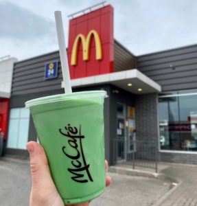 A hand holding a clear McCafe cup filled with a green beverage. In the background you can see a McDonald's restaurant with the yellow M on the outside of the building.